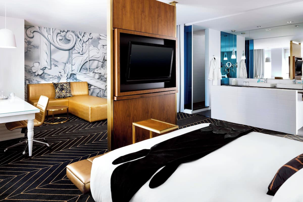 A modern hotel room with a large bed, a TV mounted on the wall, a desk with a chair, a sofa, and a small table.