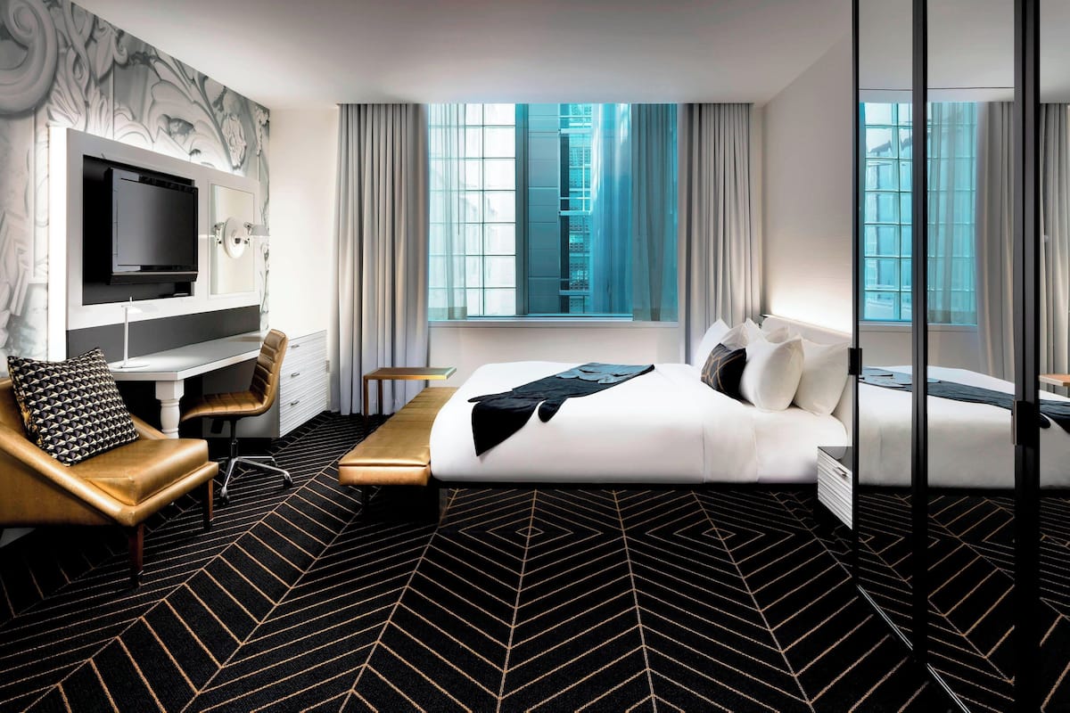 A modern hotel room with a large bed, a desk, a television, and a view of the city through large windows.