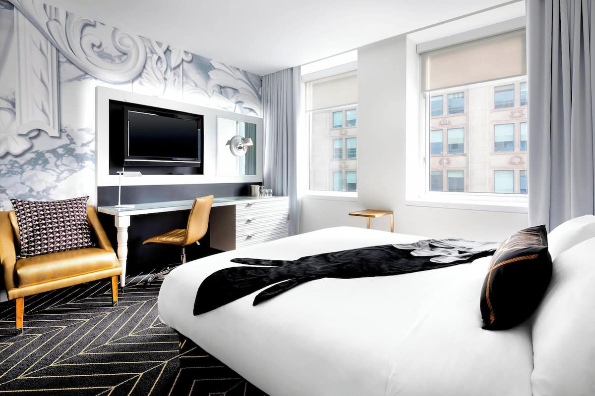 A hotel room with a large bed, a desk with a flat-screen TV, a leather armchair, and a view of the city through large windows.