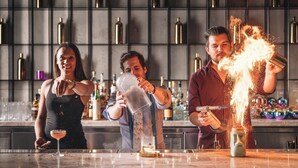 Ambiente de bar com três pessoas preparando bebidas, uma prateleira com garrafas e um balcão.