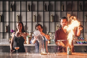 A bar setting with three individuals preparing drinks, a shelf with bottles, and a bar counter.