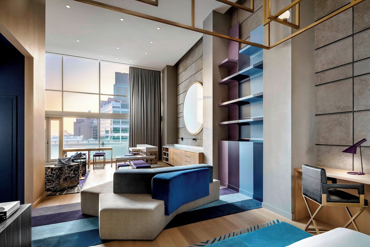A modern hotel room with a large window, a sofa, a desk, and a city view.