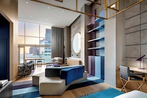 A modern hotel room with a large window, a sofa, a desk, and a city view.