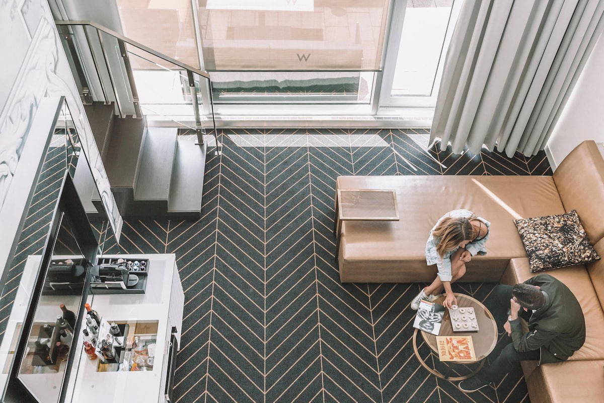 A modern hotel room with a herringbone-patterned floor, a sofa, a coffee table, and a view of the outdoors through a large window.