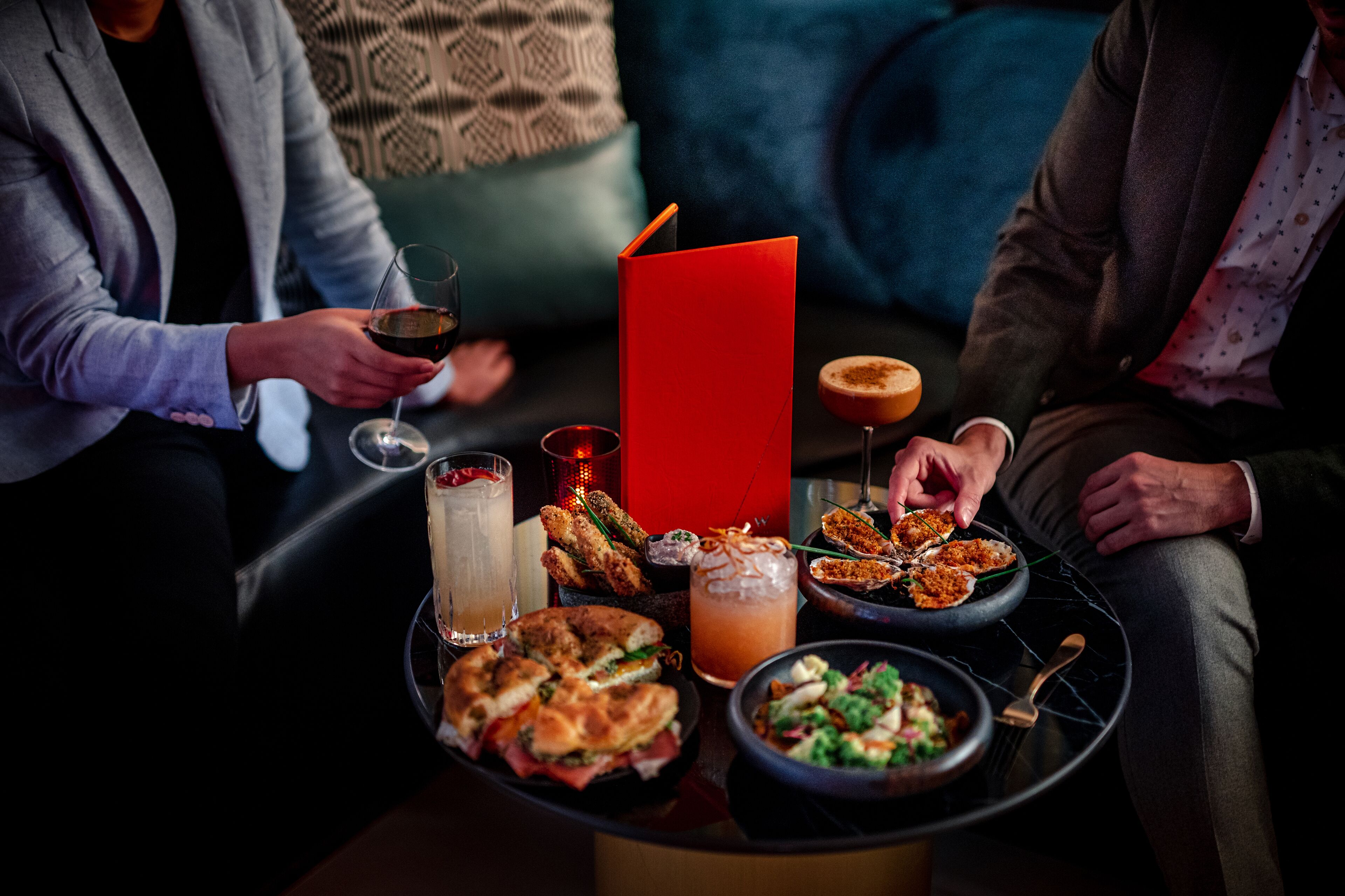 A dining table with various dishes and drinks, surrounded by people seated on a couch.