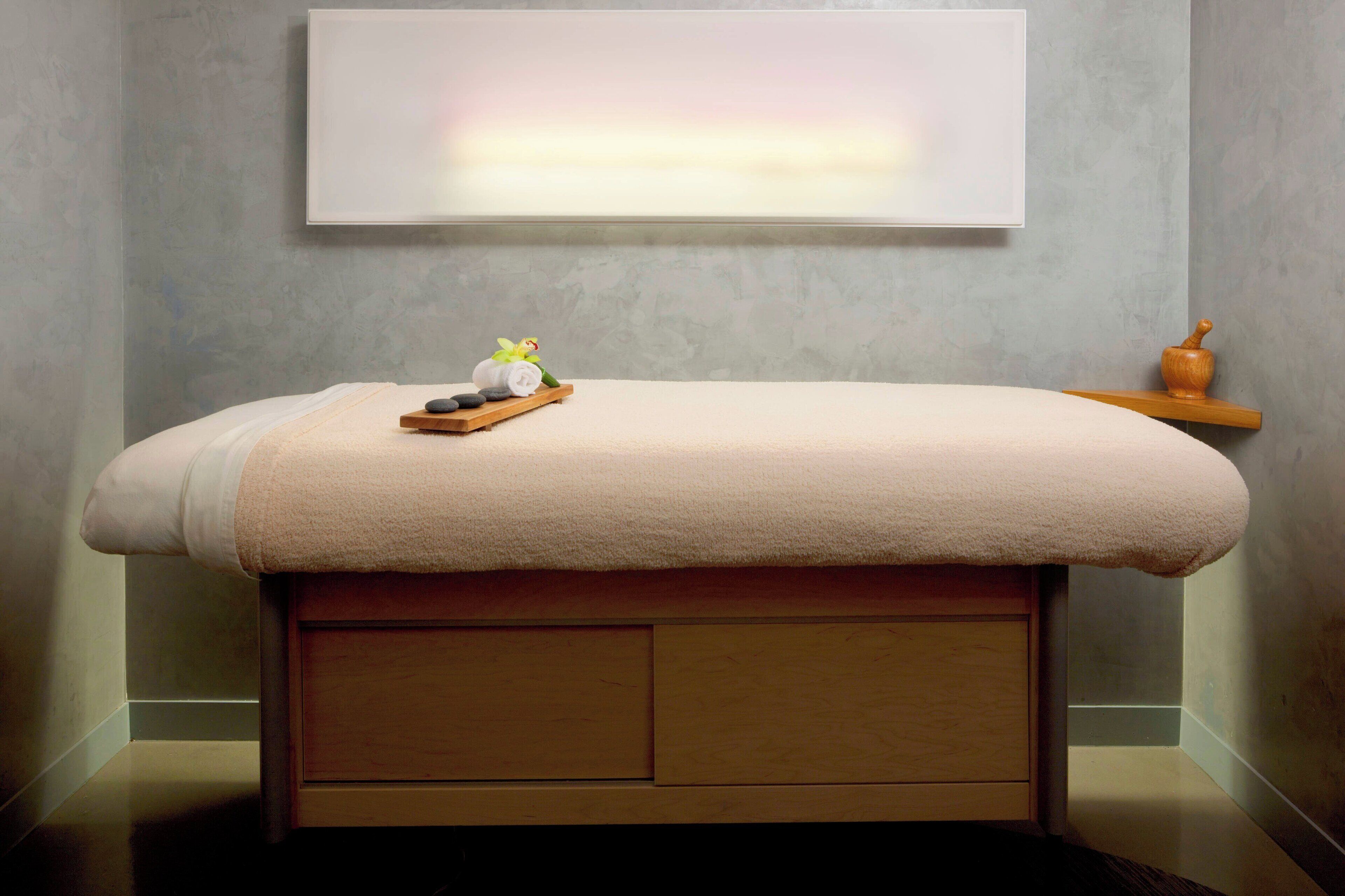 A spa treatment room with a massage table, a wooden shelf with a stone, a rolled towel, and a small vase.