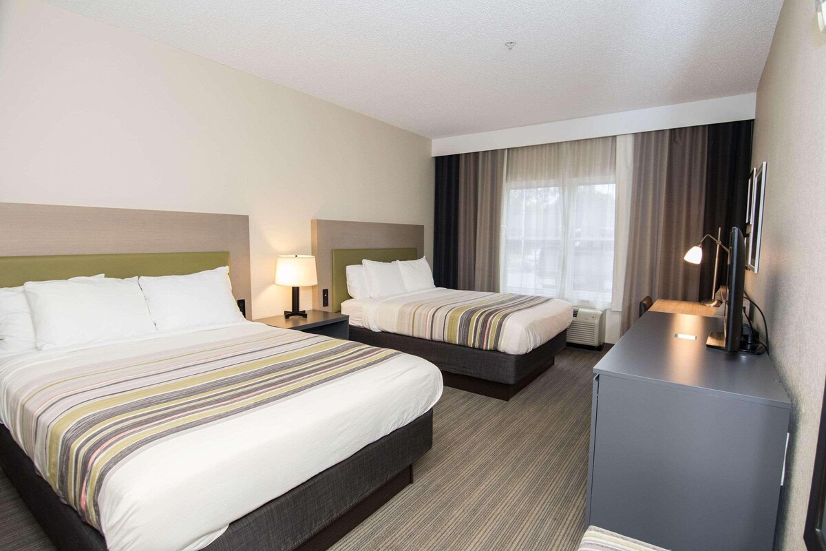 A hotel room with two beds, a desk, a chair, a lamp, and a large window with curtains.