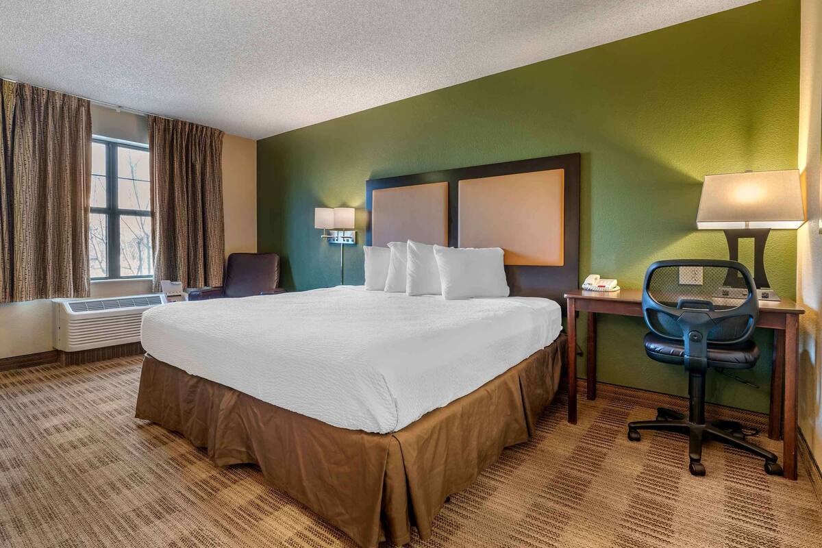 Habitación de hotel con una cama grande, un escritorio con una silla, una lámpara y una ventana con cortinas.