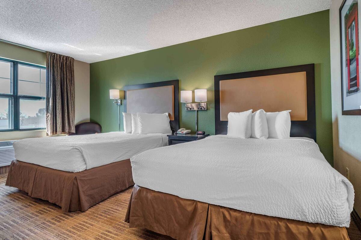 Habitación de hotel con dos camas, un escritorio con lámpara, una silla y un ventanal con cortinas.