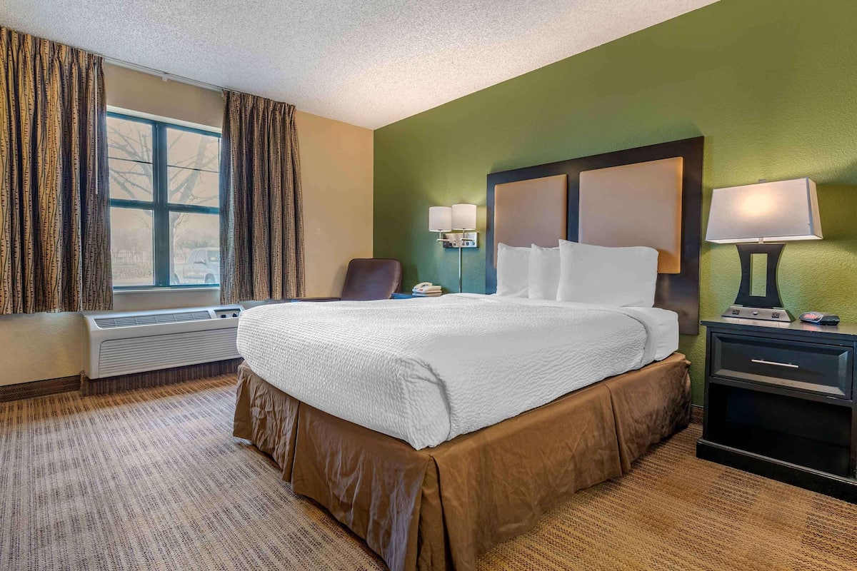 Habitación de hotel con una cama grande, una silla, un escritorio con lámpara y una ventana con cortinas.