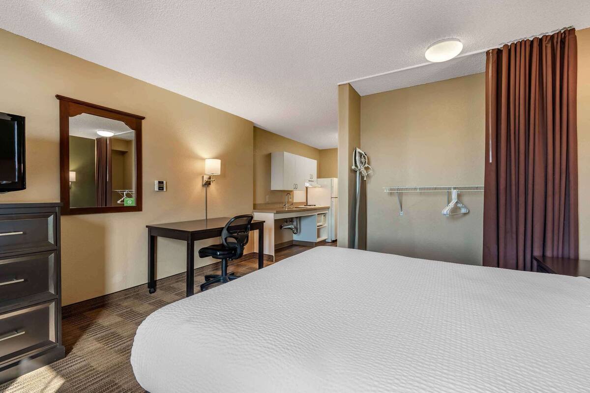 Habitación de hotel con una cama grande, un escritorio con silla, un espejo, un televisor y un baño con ducha, además de una cortina.