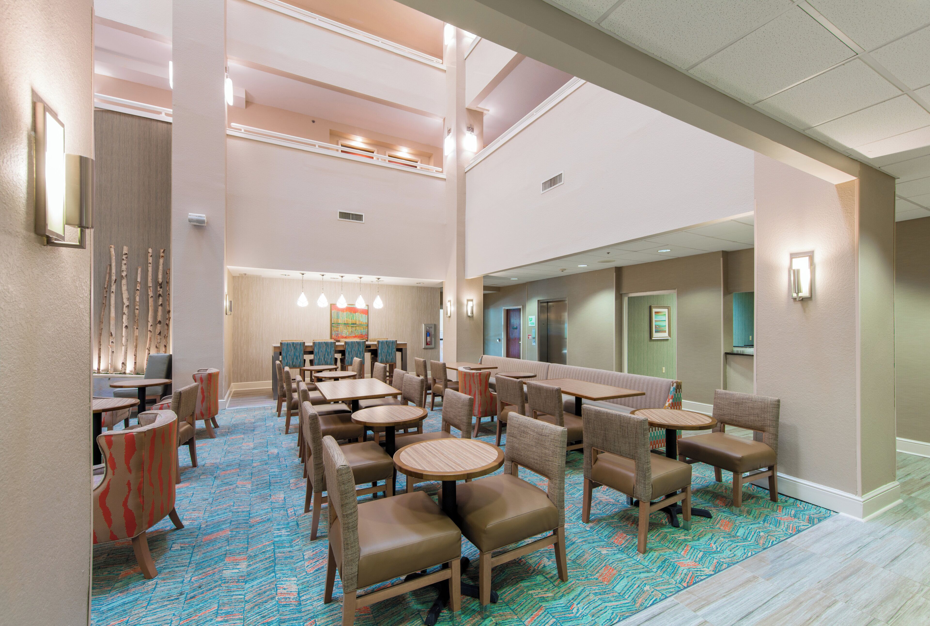 A hotel lobby with multiple seating areas, including round tables and booths, and a high ceiling with recessed lighting.