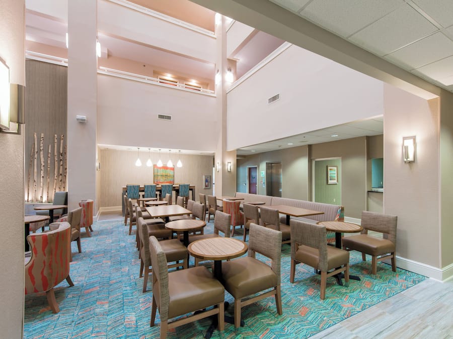 Un hall de hotel con varias zonas de estar, incluyendo mesas redondas y cabinas, y un techo alto con iluminación empotrada.