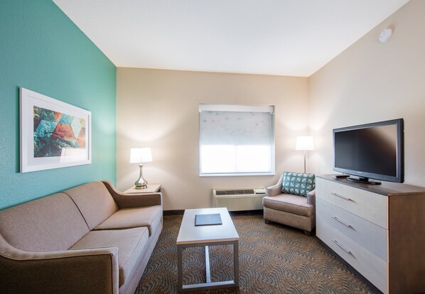 A hotel room with a beige sofa, a small table with a book, a TV on a wooden cabinet, and a window with blinds. - Holiday Inn & Suites Lake City by IHG (Lake City)