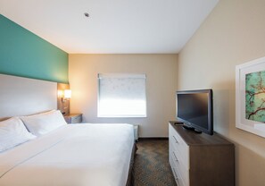 A hotel room with a bed, a television on a wooden dresser, a nightstand with a lamp, and a window with blinds. - Holiday Inn & Suites Lake City by IHG (Lake City)