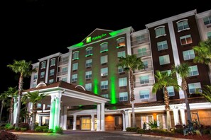 Un hotel de varios pisos con una entrada destacada, palmeras y un letrero iluminado en verde que indica el nombre.