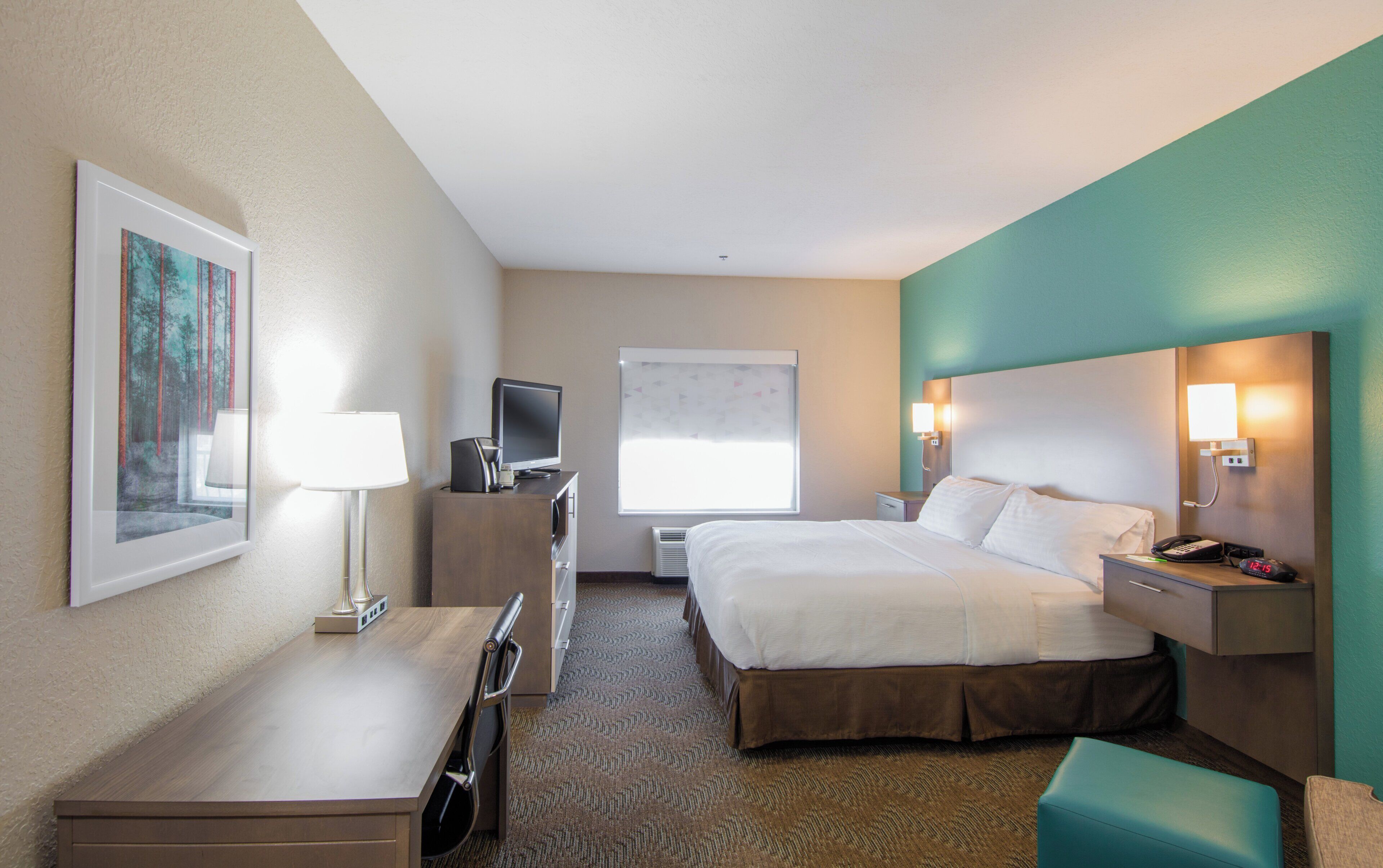 A hotel room with a bed, desk, chair, television, bedside table, lamp, and a window with blinds.