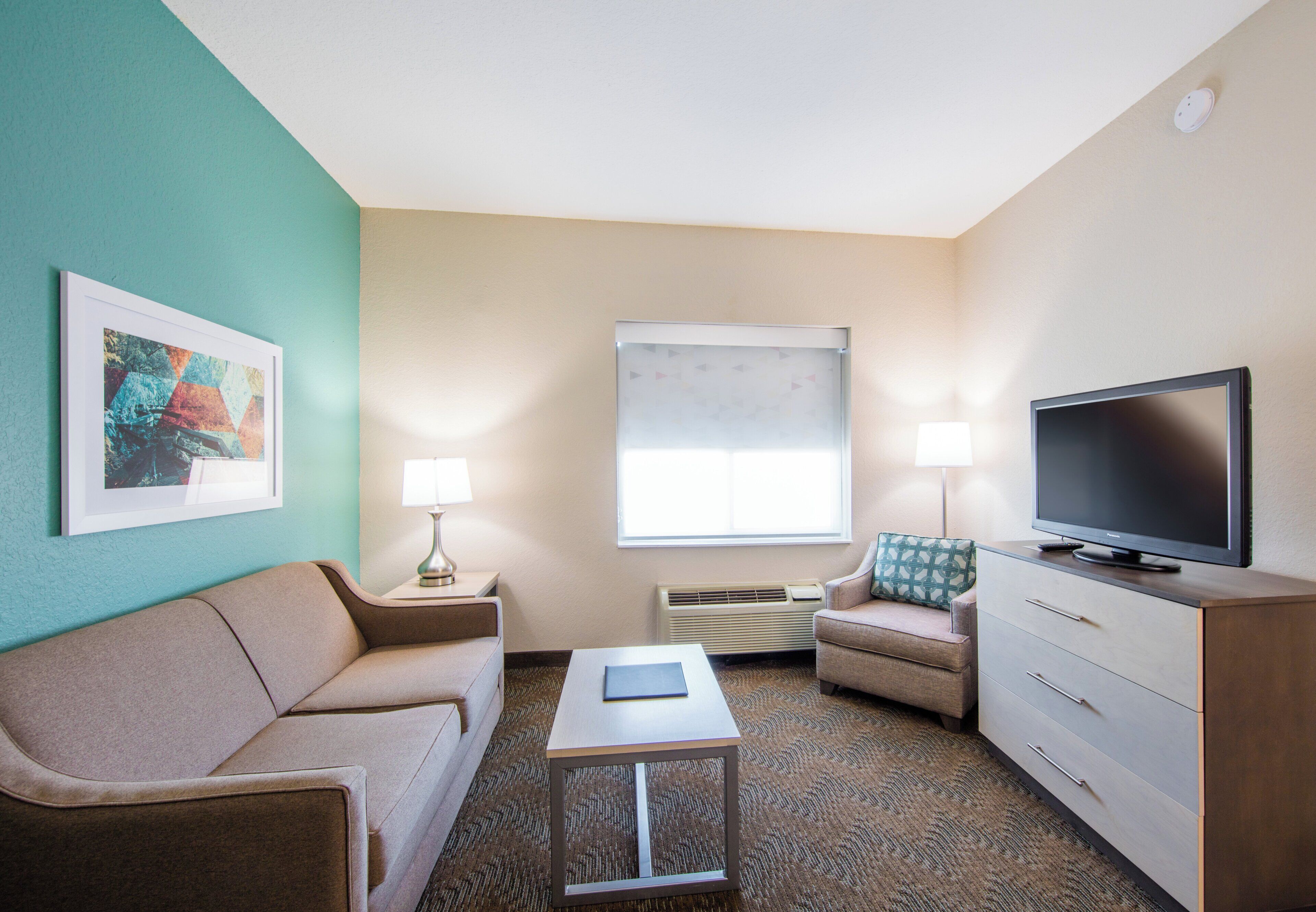 Habitación de hotel con sofá, mesa de centro, televisor, lámpara y ventana.