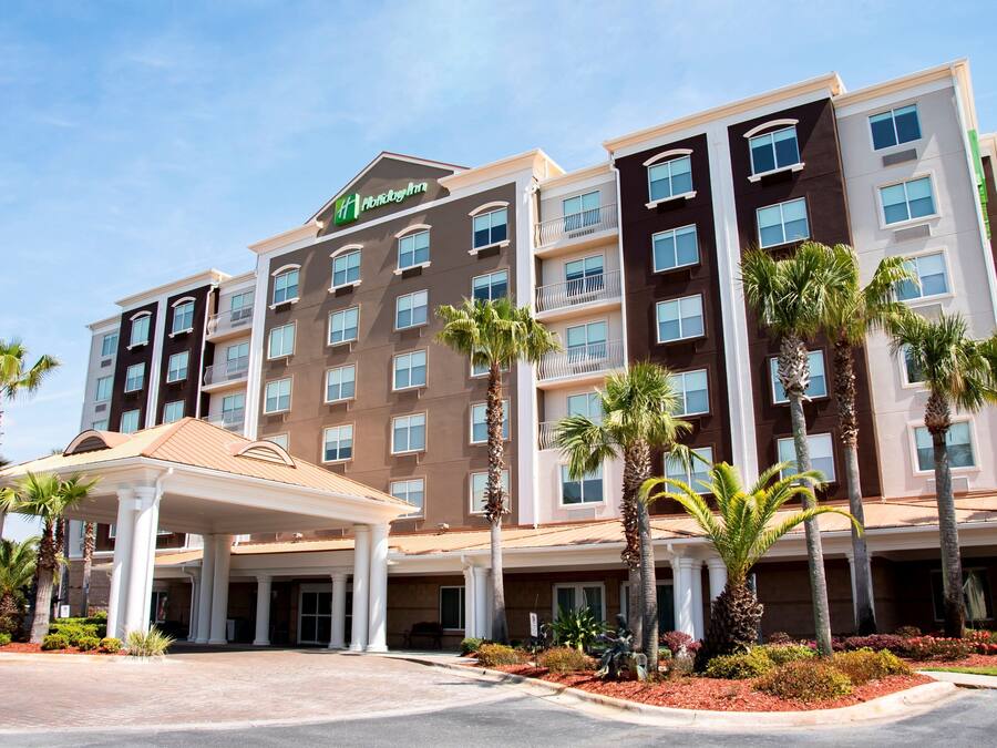A multi-story hotel with a central entrance, palm trees, and a covered walkway.