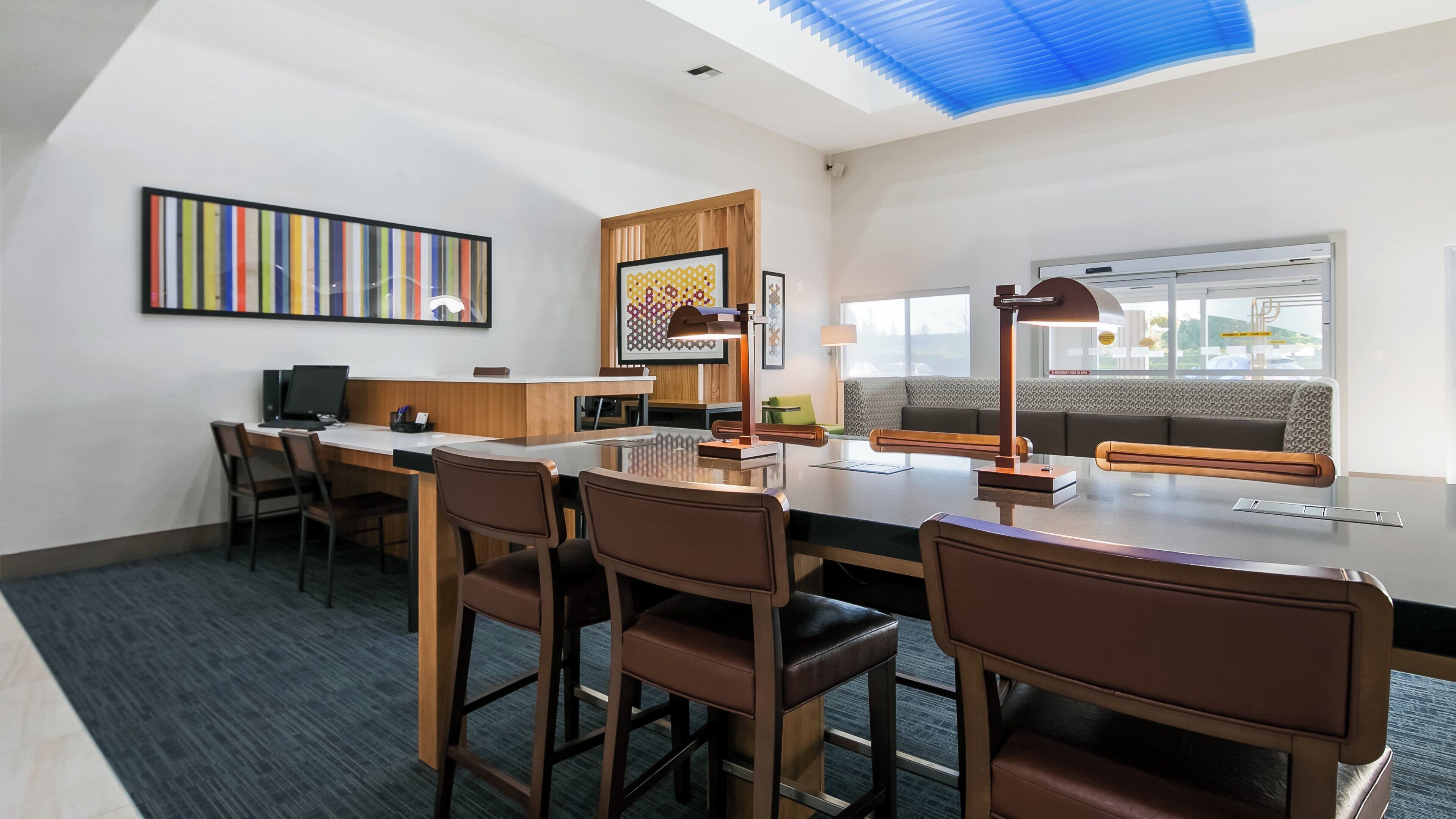 A modern hotel room with a large desk, multiple chairs, a sofa, a ceiling light, and a window with a view.