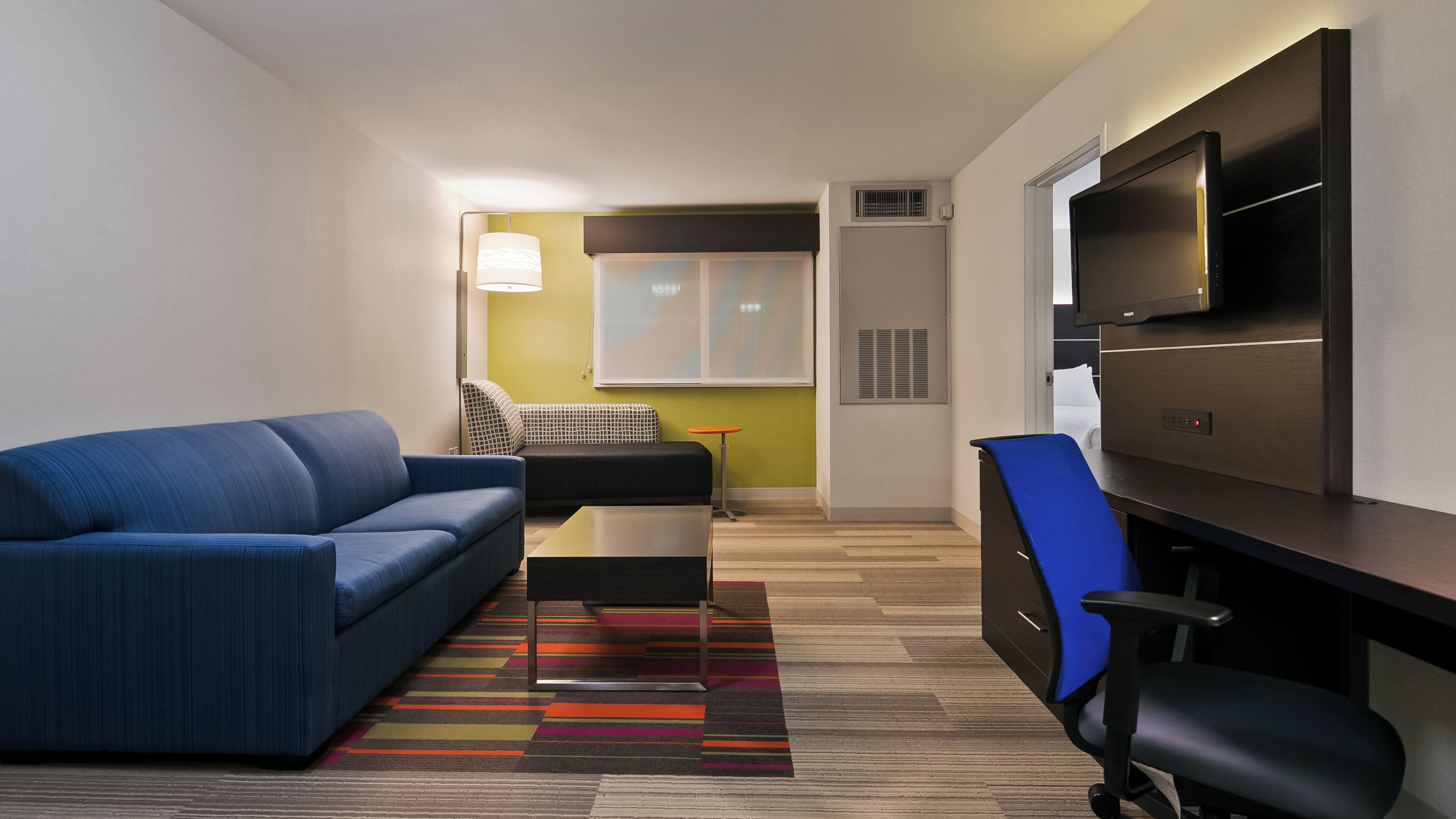 Et moderne hotelværelse med en blå sofa, et lille bord, et skrivebord med stol, et fjernsyn og en farverig stribet måtte.