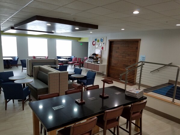 A modern cafe with a variety of seating options including tables, chairs, and a sofa. There is a coffee station with a counter and a sign that reads "Smart Roasters." A staircase with a metal railing is visible in the background. - Holiday Inn Express & Suites Everett by IHG (Everett)