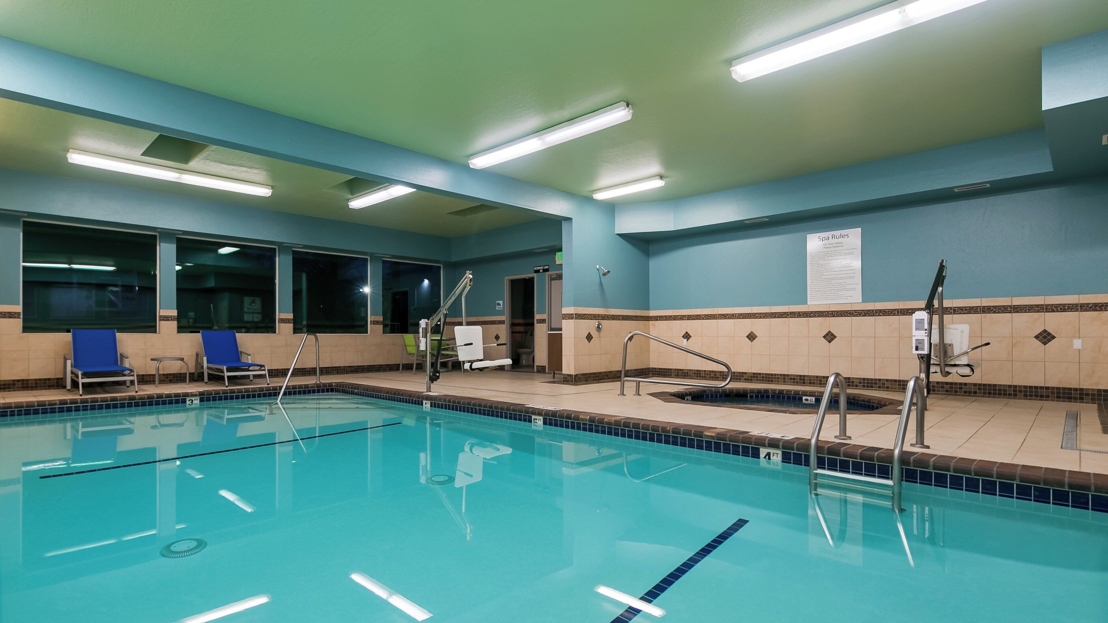 A swimming pool with clear lane dividers, a changing room with blue chairs, and a sign indicating spa rules.