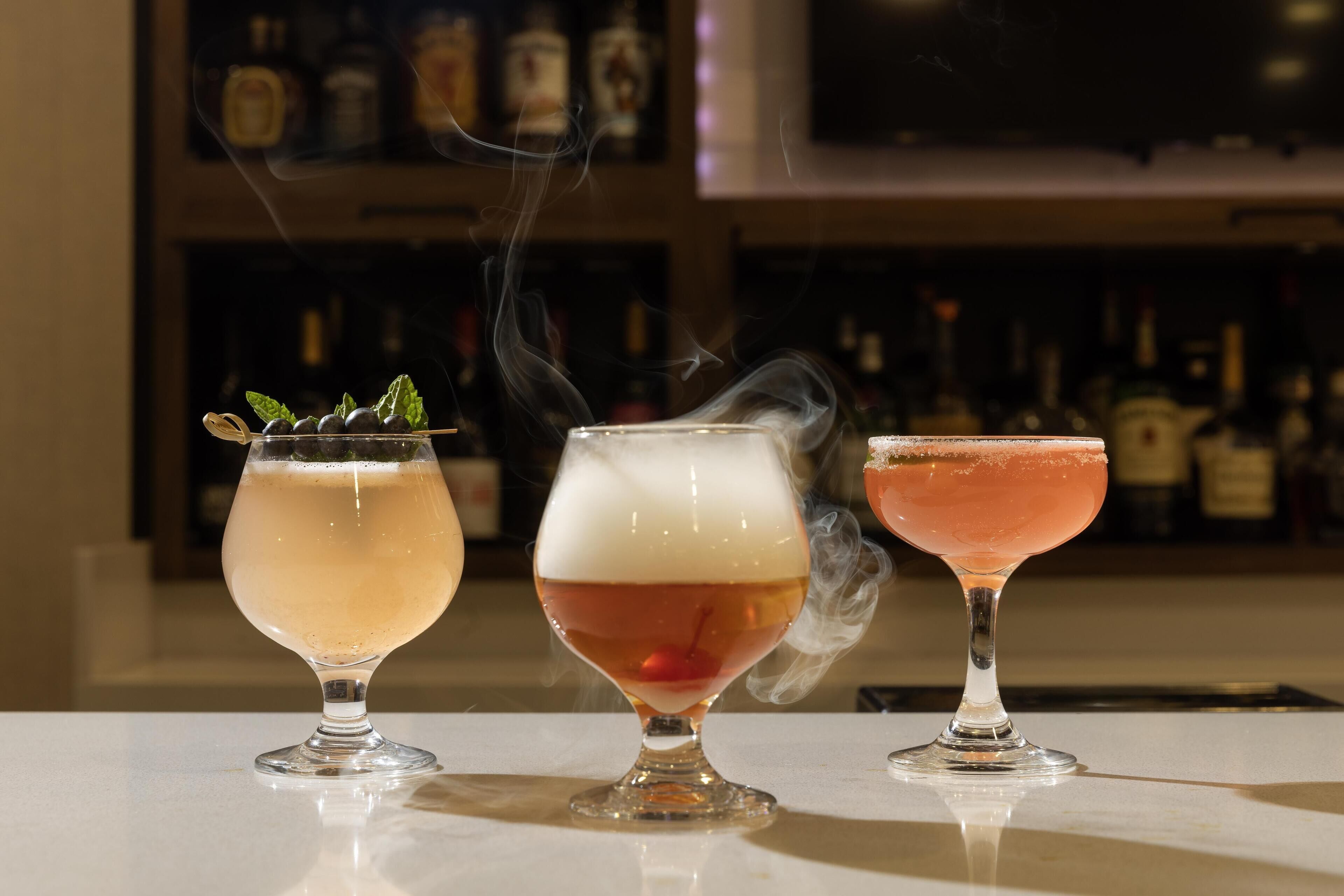 Three cocktails on a bar counter with a blurred background of bottles.