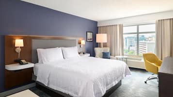 A hotel room with a large bed, bedside lamps, a chair, a desk, and a view of the city through the window.