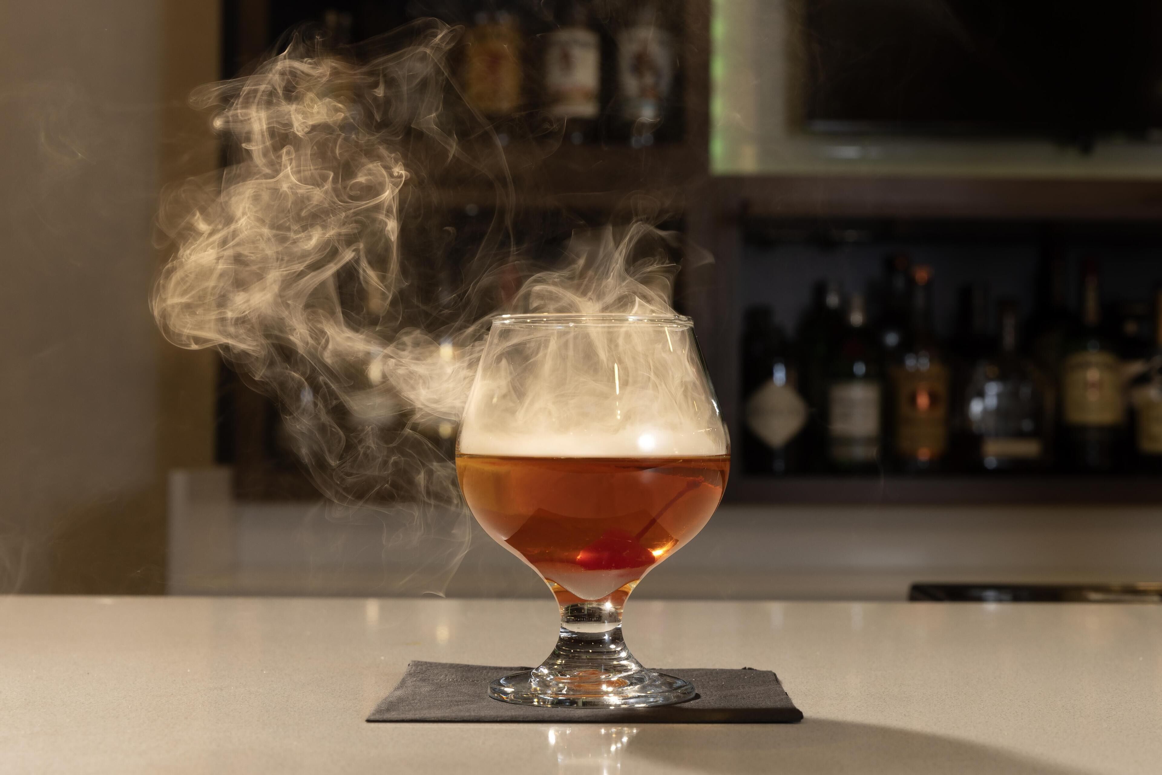 Un vaso de líquido color ámbar con espuma, que emite vapor, sobre un posavasos de bar.