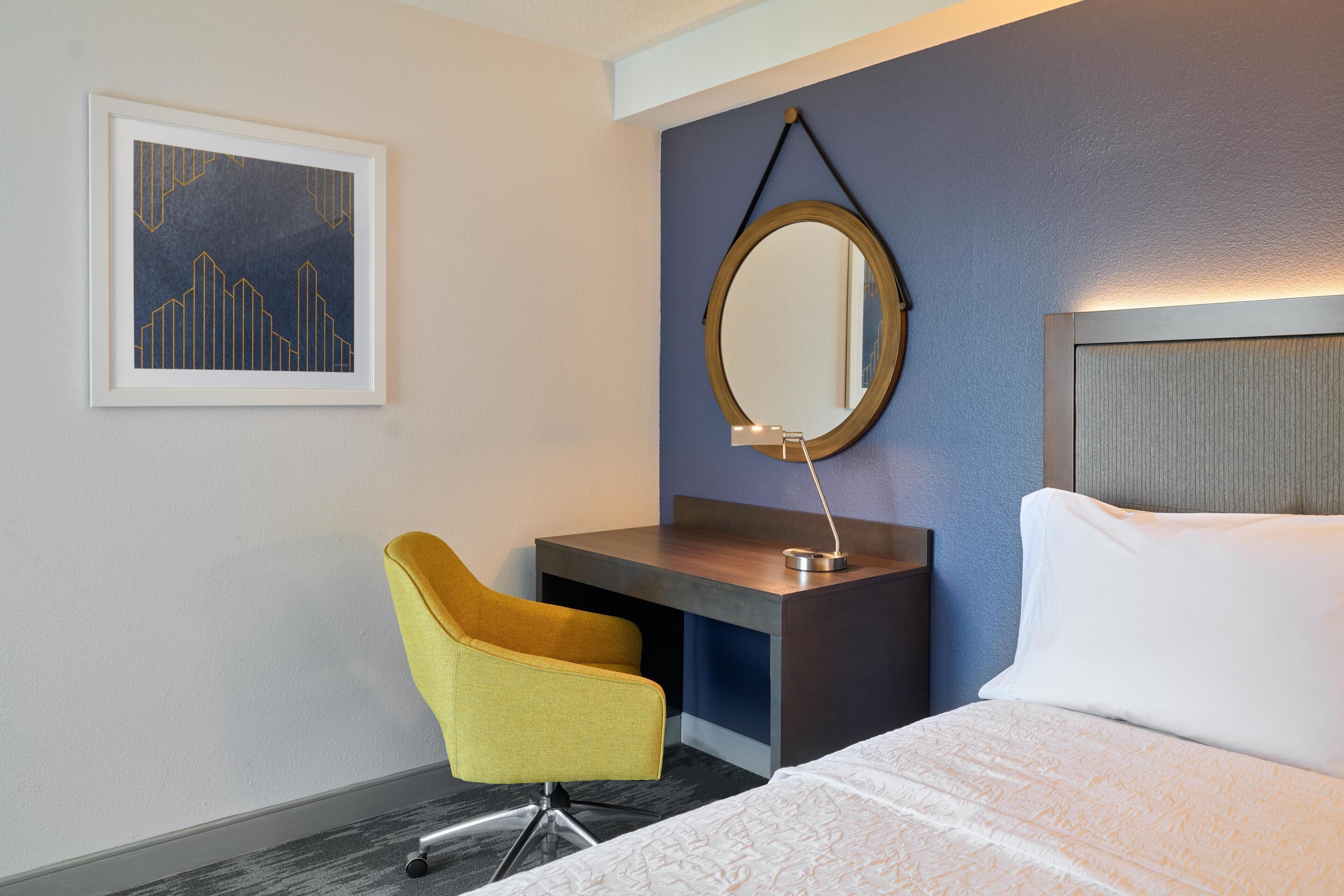 Habitación de hotel con cama, escritorio con silla, un cuadro en la pared y un espejo redondo.