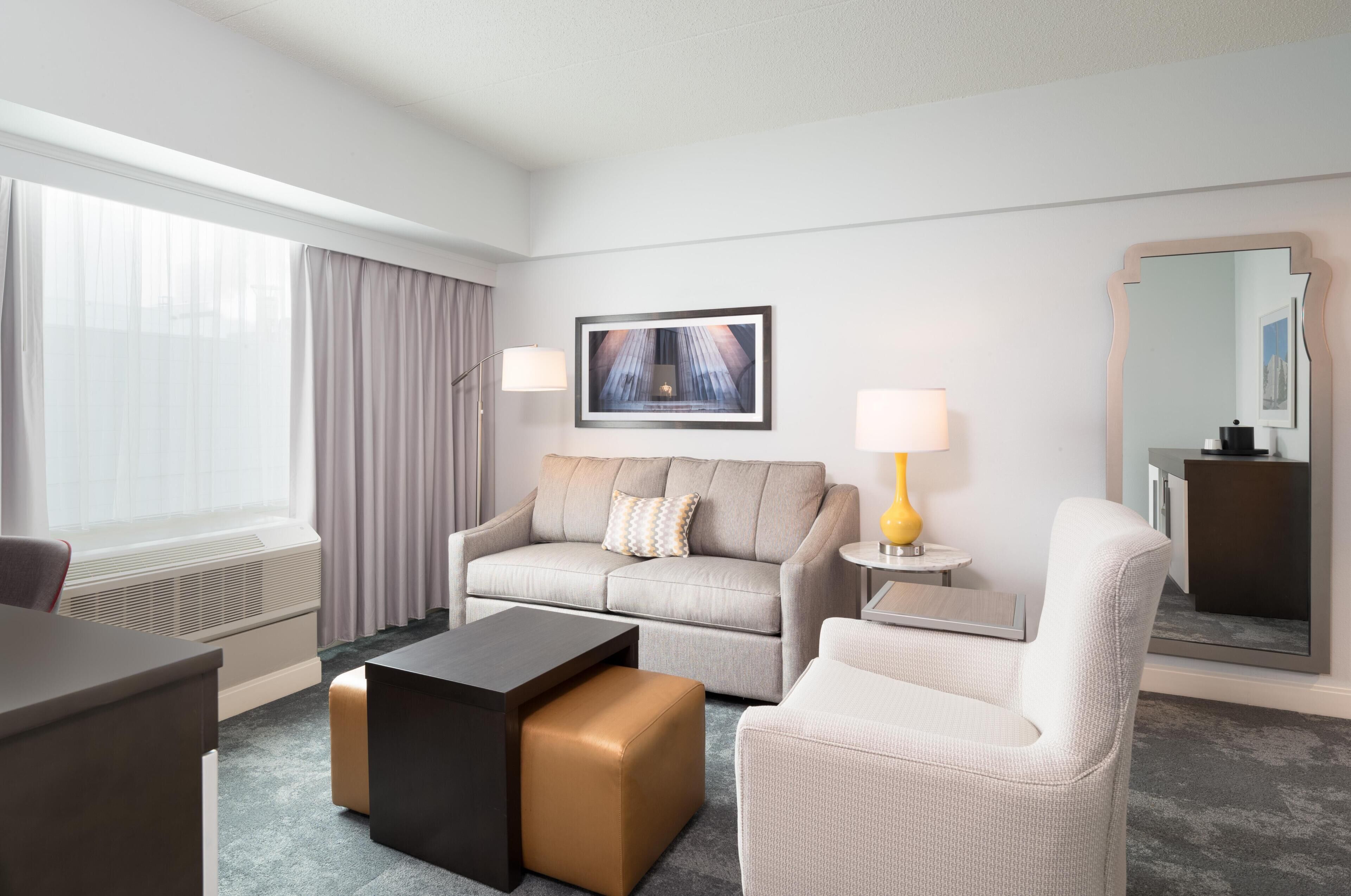 A hotel room with a sofa, armchair, coffee table, side table with lamp, and a large mirror.