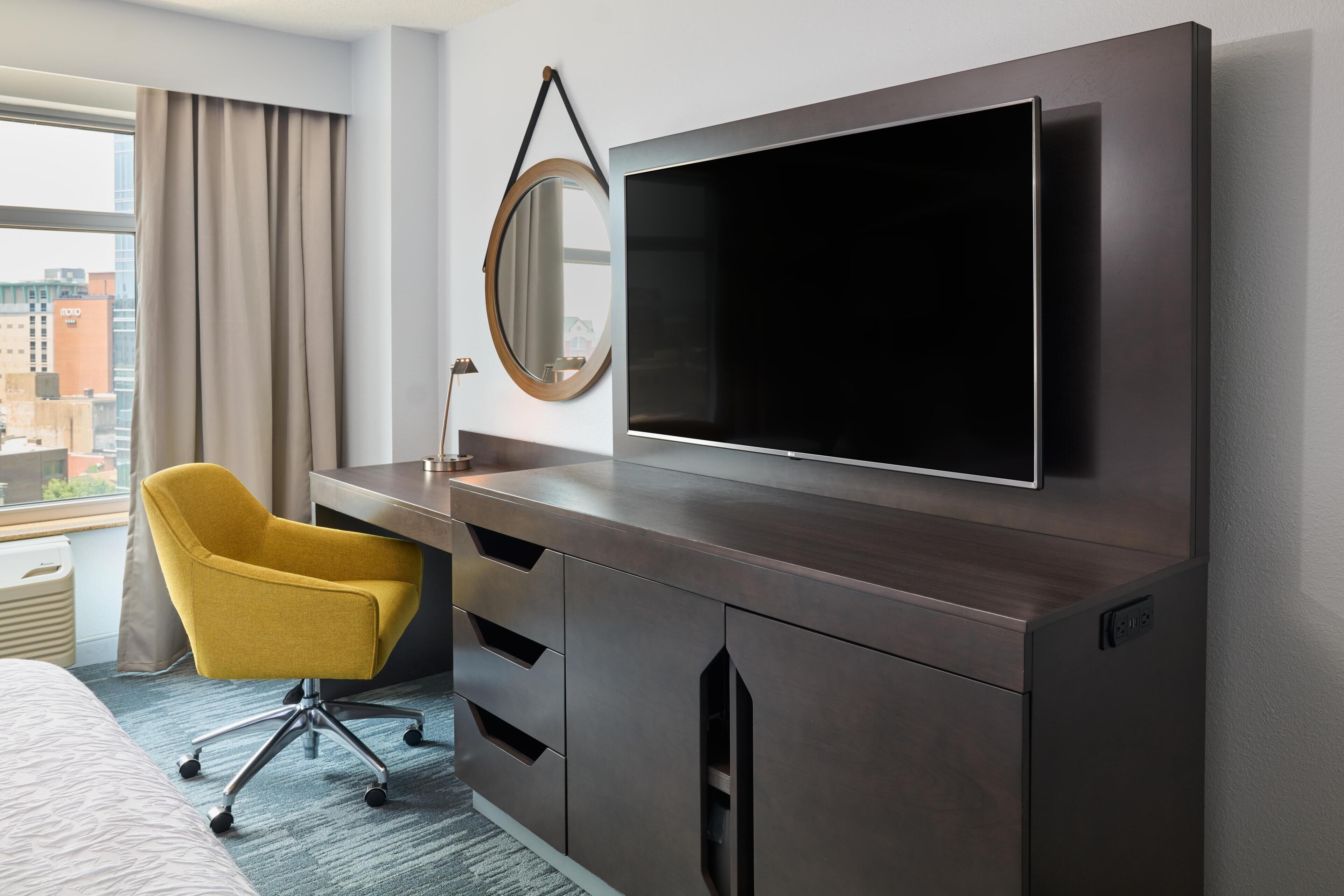 A hotel room with a large flat-screen TV mounted on a dark wooden cabinet, a yellow office chair, a round mirror, and a view of the city through a large window.