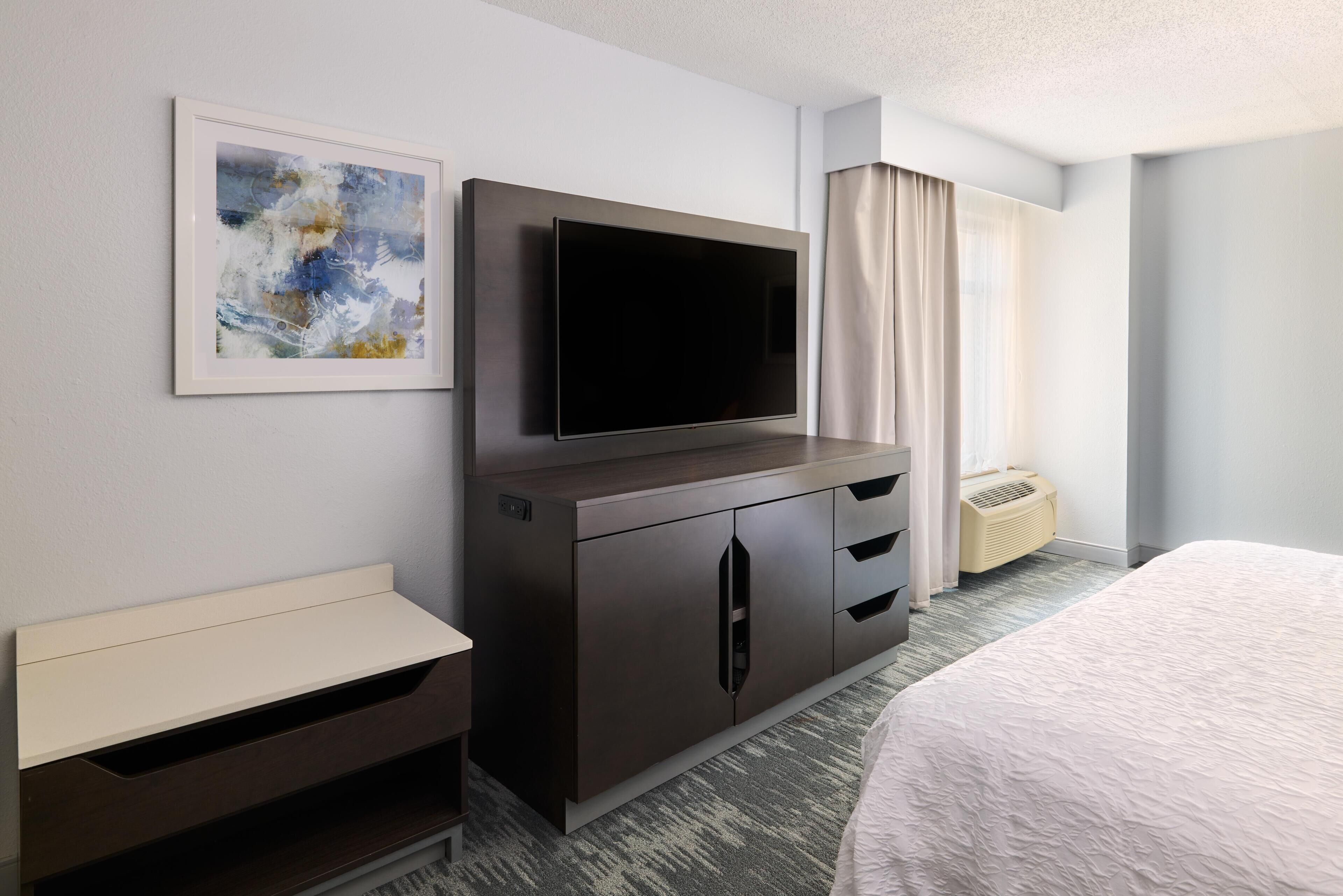 A hotel room with a flat-screen TV, a bed, a small desk, a framed abstract painting, and a window with curtains.
