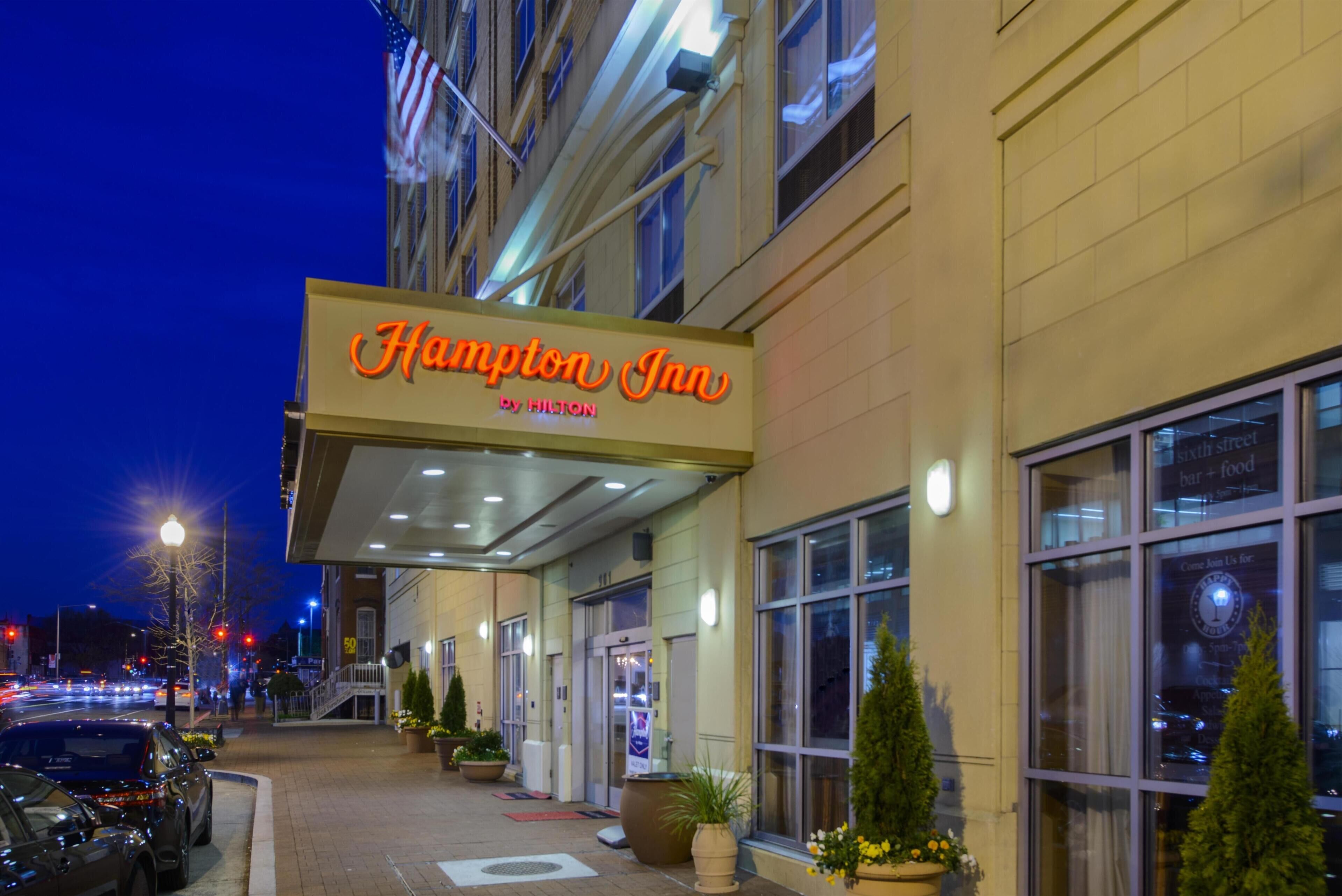 A hotel entrance with a neon sign, a covered walkway, and potted plants.