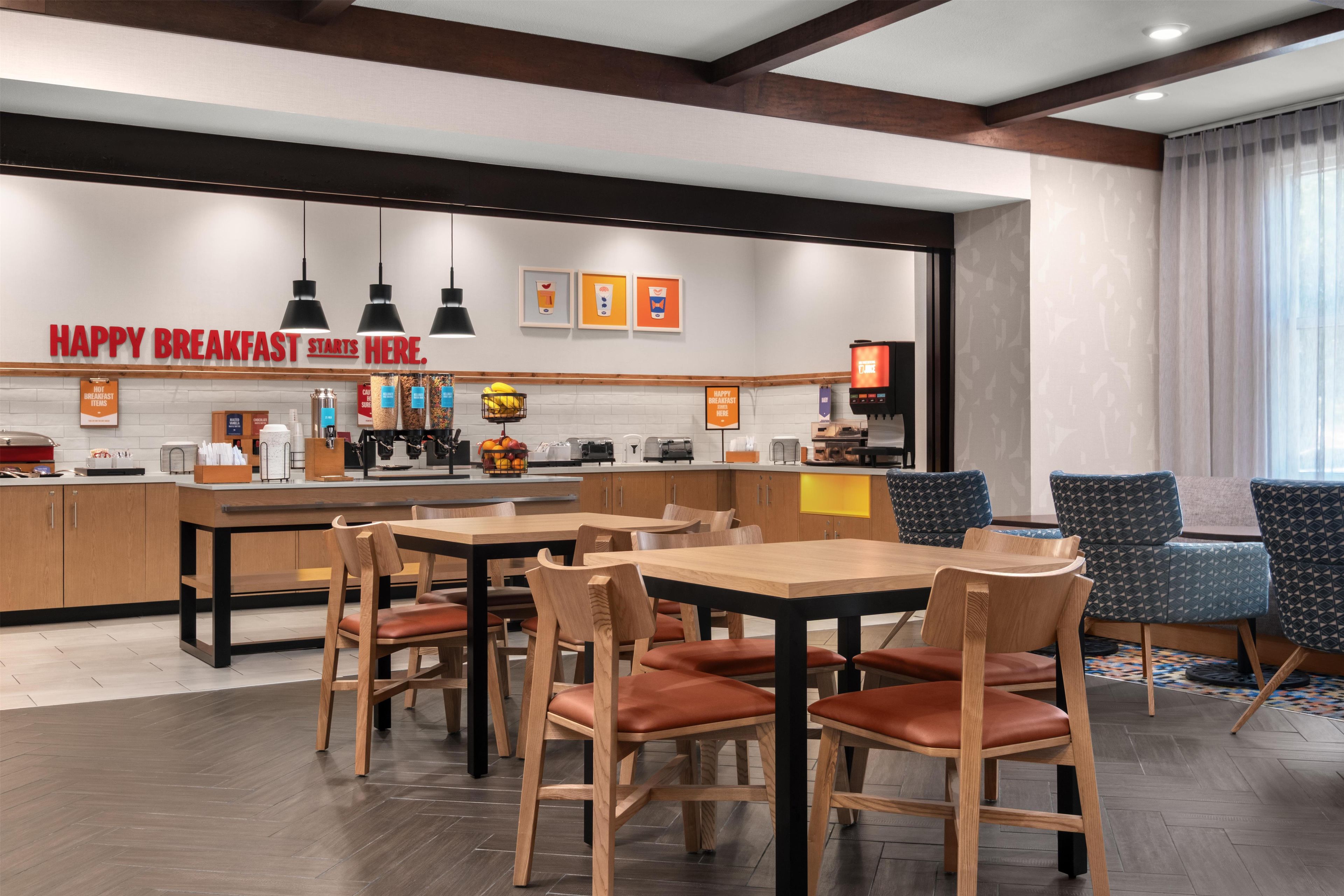 A modern breakfast area with wooden tables and chairs, a coffee machine, and a sign that reads 'HAPPY BREAKFAST STARTS HERE.'