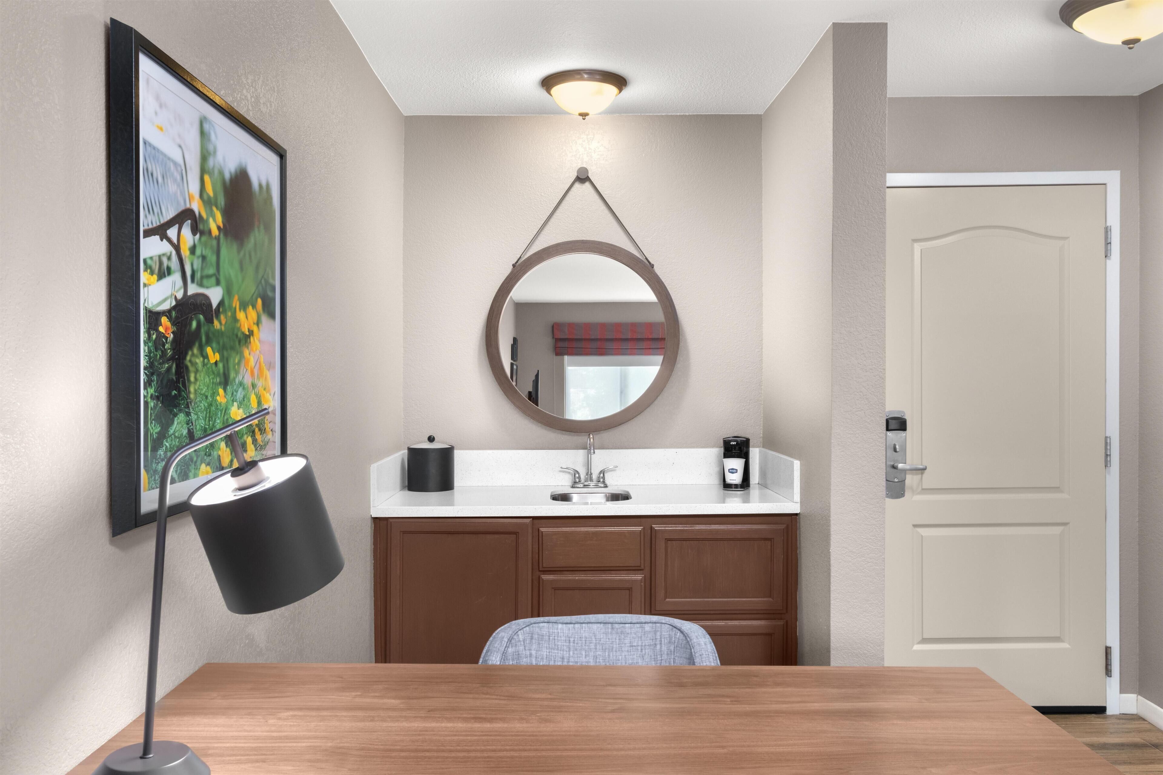 Habitación de hotel con un escritorio de madera, un espejo redondo, un cuadro enmarcado en la pared, un lavamanos con cafetera y una puerta.