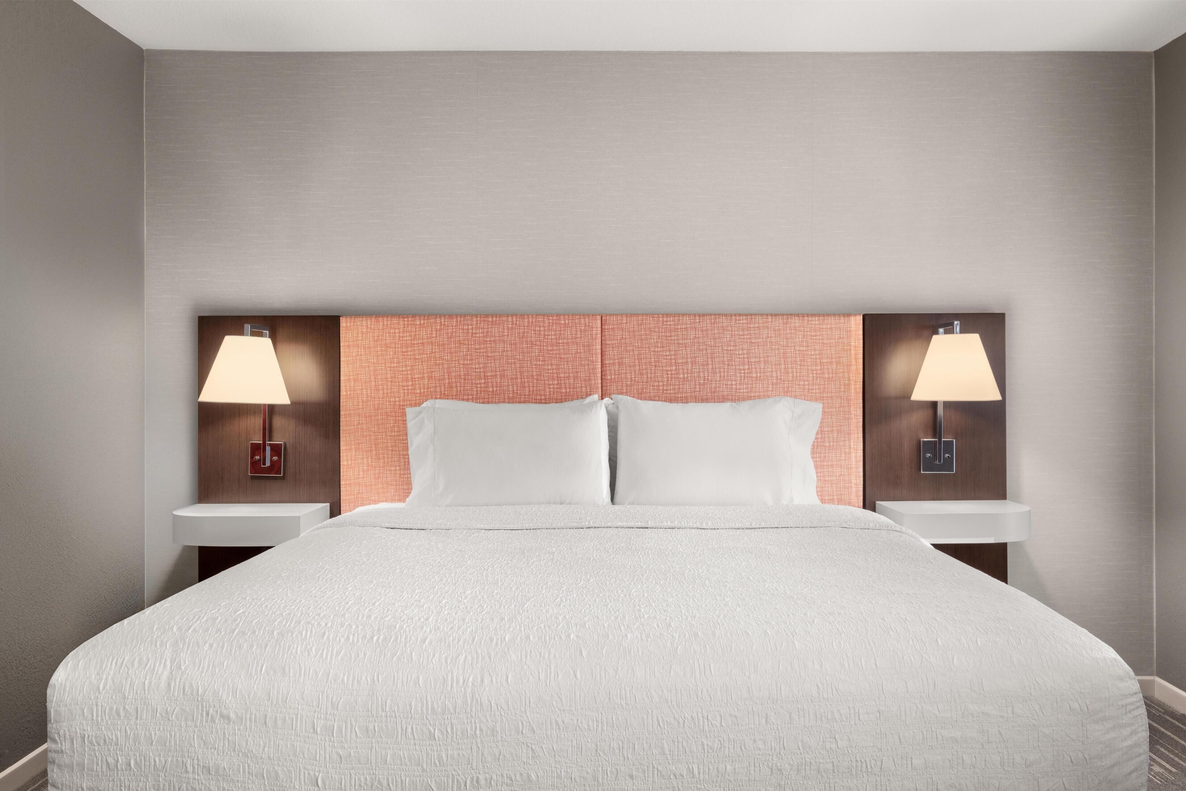 A hotel room with a large bed, two bedside tables with lamps, a headboard, and a plain wall.