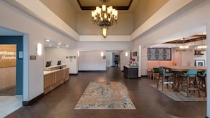 A hotel lobby with a reception desk, a chandelier, a dining area with chairs, and a kitchen in the background. - Hampton Inn & Suites Fresno (Fresno)
