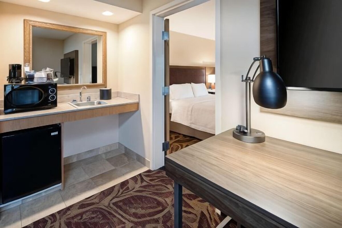A hotel room with a bed, a desk, a desk lamp, a TV, a microwave, and a sink.
