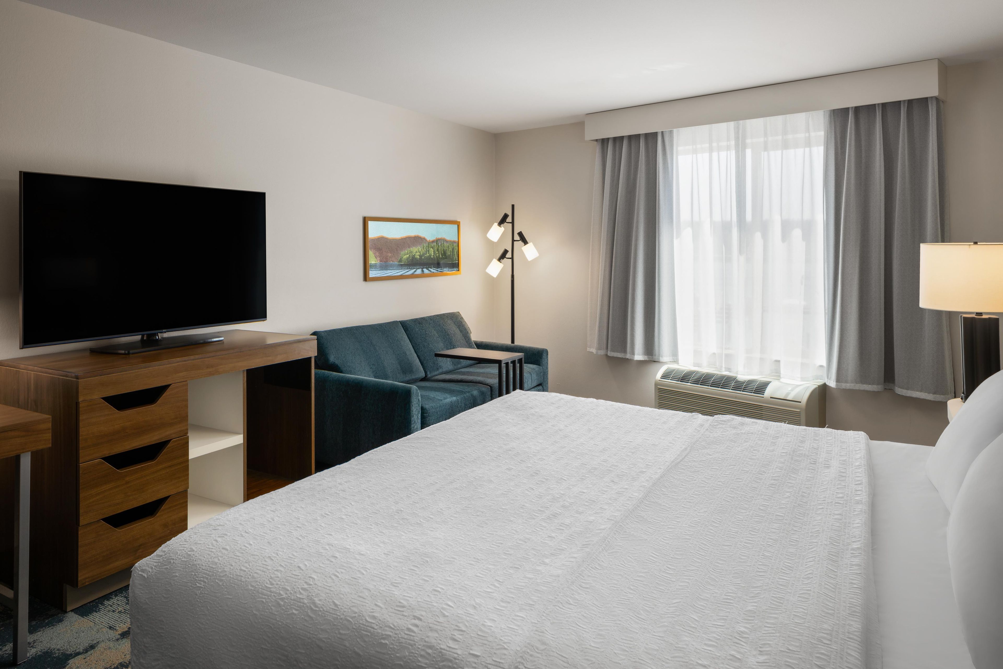 A hotel room with a bed, a desk with a TV, a chair, a picture on the wall, a floor lamp, and a window with curtains.