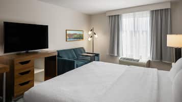 A hotel room with a bed, a desk with a TV, a chair, a picture on the wall, a floor lamp, and a window with curtains.