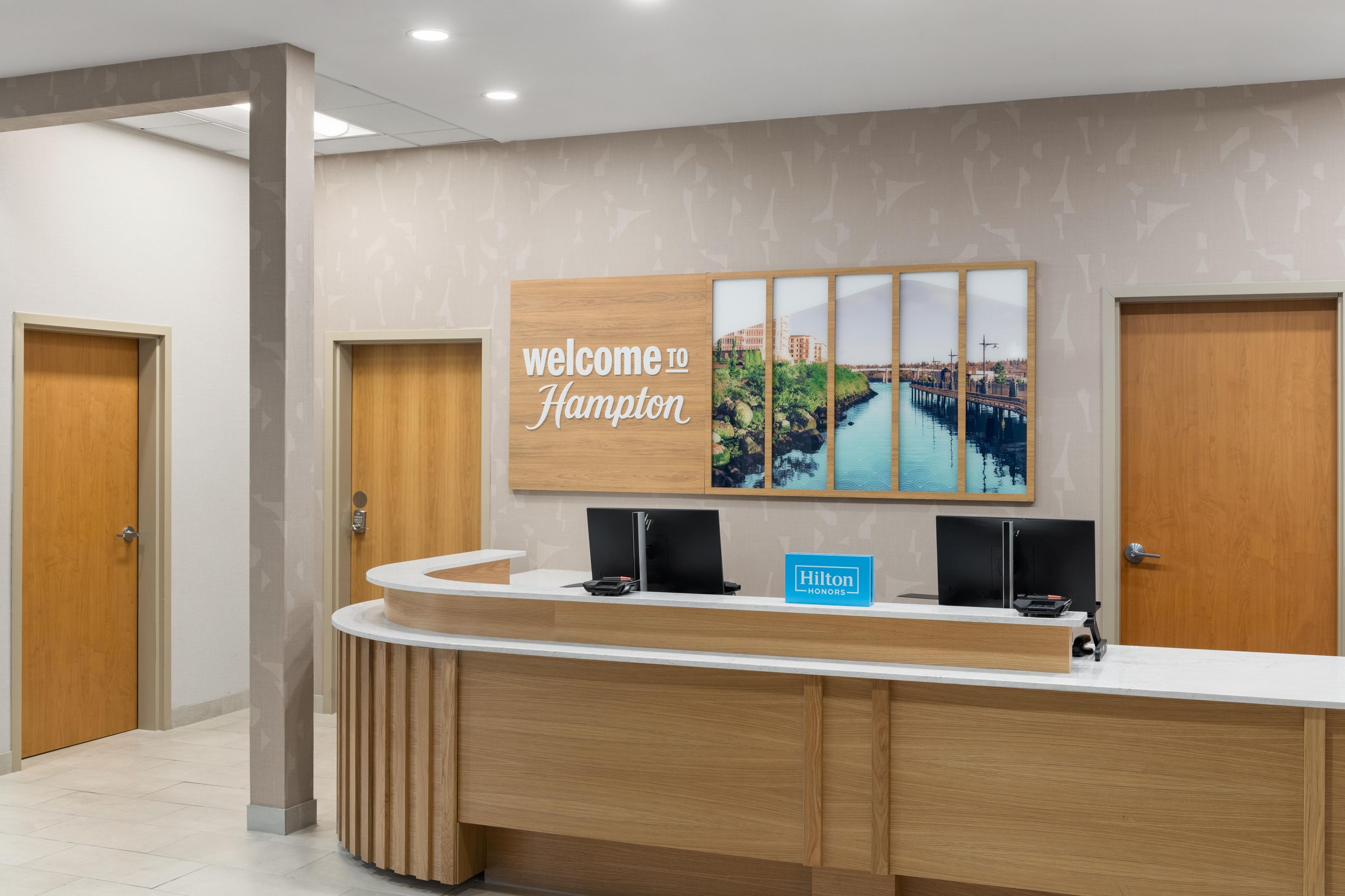 A hotel reception desk with a 'welcome to Hampton' sign, a blue 'Hilton Honors' sign, and a cityscape artwork.