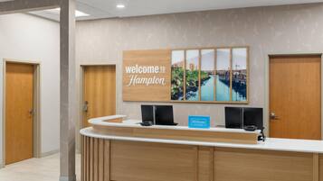 A hotel reception desk with a 'welcome to Hampton' sign, a blue 'Hilton Honors' sign, and a cityscape artwork.