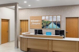 A hotel reception desk with a 'welcome to Hampton' sign, a blue 'Hilton Honors' sign, and a cityscape artwork.