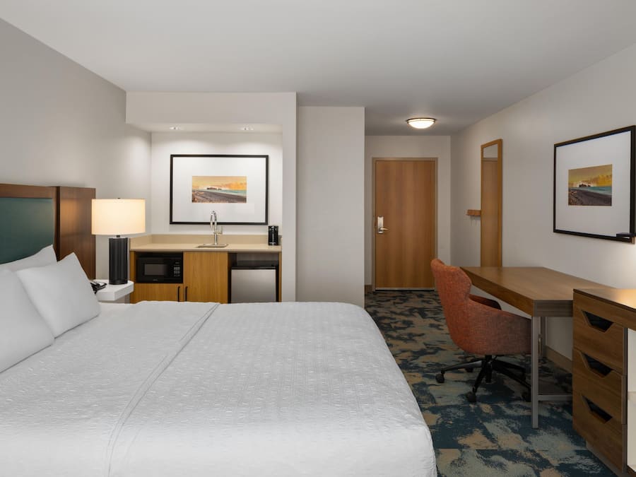 Ein Hotelzimmer mit einem großen Bett, einem Schreibtisch mit Stuhl, einer Mikrowelle, einer Kaffeemaschine und einem gerahmten Bild an der Wand.