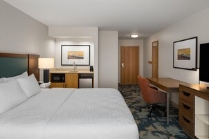 Habitación de hotel con una cama grande, un escritorio con silla, microondas, cafetera y un cuadro en la pared.