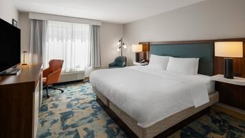 A hotel room with a large bed, a desk with a flat-screen TV, a chair, a bedside table with a lamp, and a window with curtains.