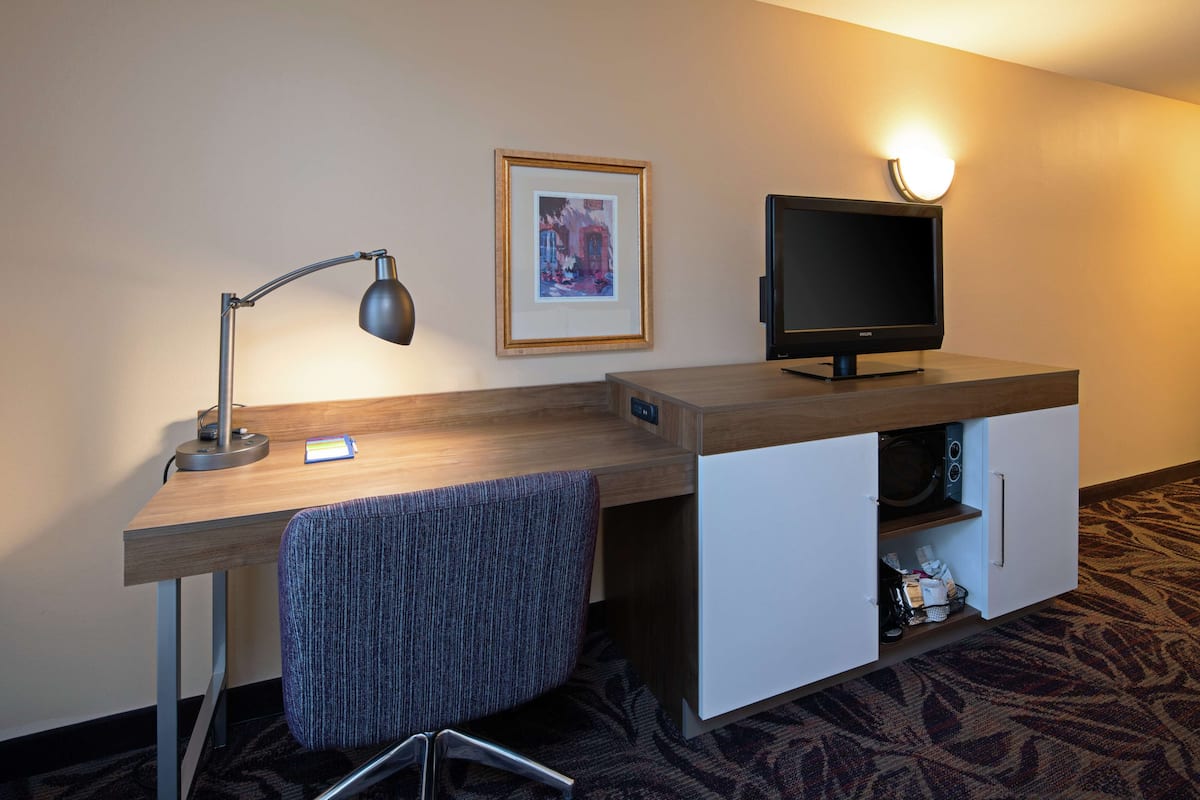 A hotel room with a desk, a chair, a television, a framed picture on the wall, and a microwave in a cabinet.