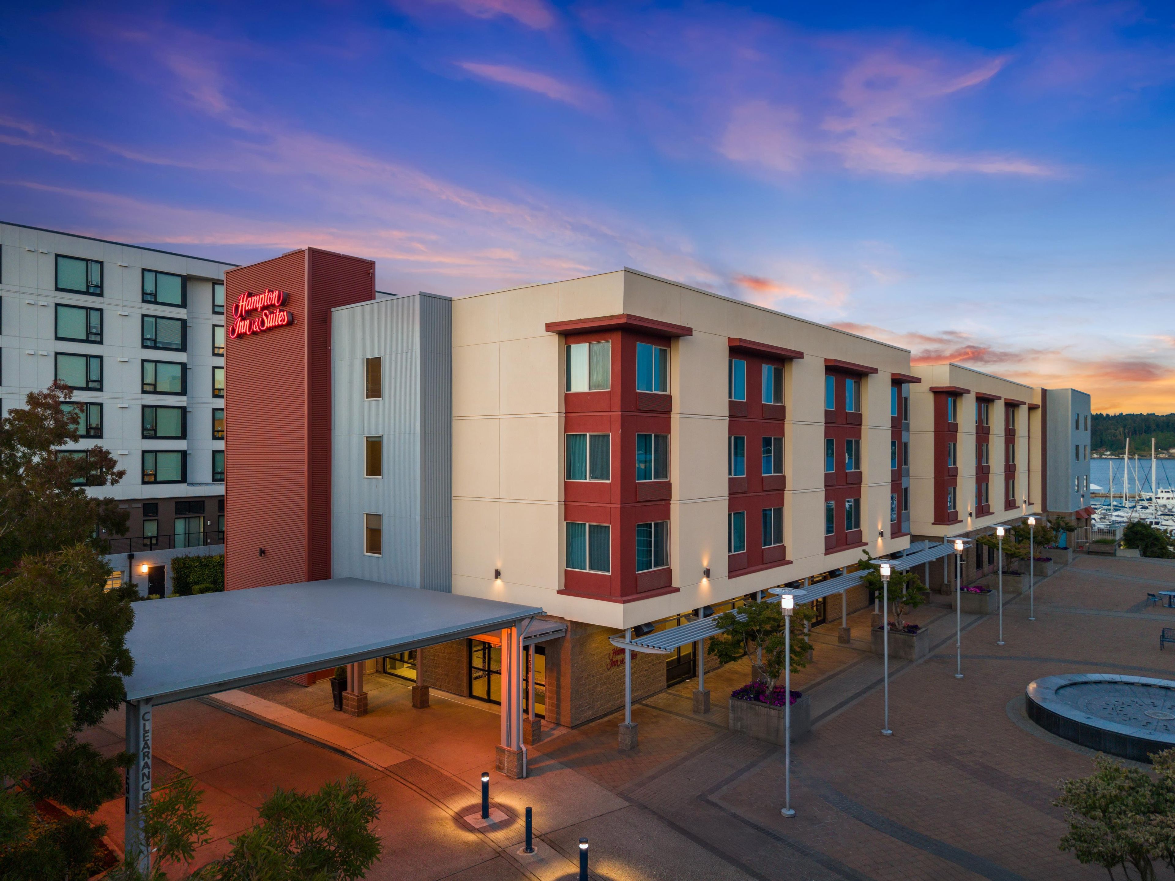 Un hotel con diseño moderno, que destaca por su fachada roja y blanca, un área exterior cubierta y vistas a la marina al fondo.